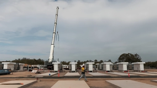 Rows of rectangular battery enclosures sit on concrete pads as a worker walks past in the foreground