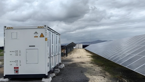 White battery storage container beside a dirt road leading toward rows of solar panels under a cloudy sky