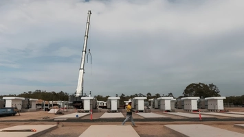 Rows of rectangular battery enclosures sit on concrete pads as a worker walks past in the foreground