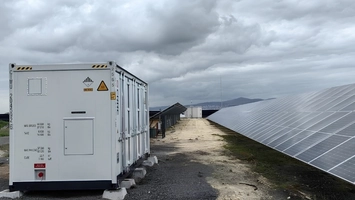 White battery storage container beside a dirt road leading toward rows of solar panels under a cloudy sky